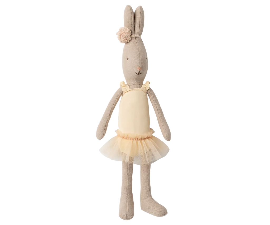 The Maileg Rabbit, Size 1 - Classic - Ballet Suit and Skirt - Cream is posed like a ballet dancer, wearing a yellow headband with a pompom and a yellow tulle tutu dress against a neutral striped background.