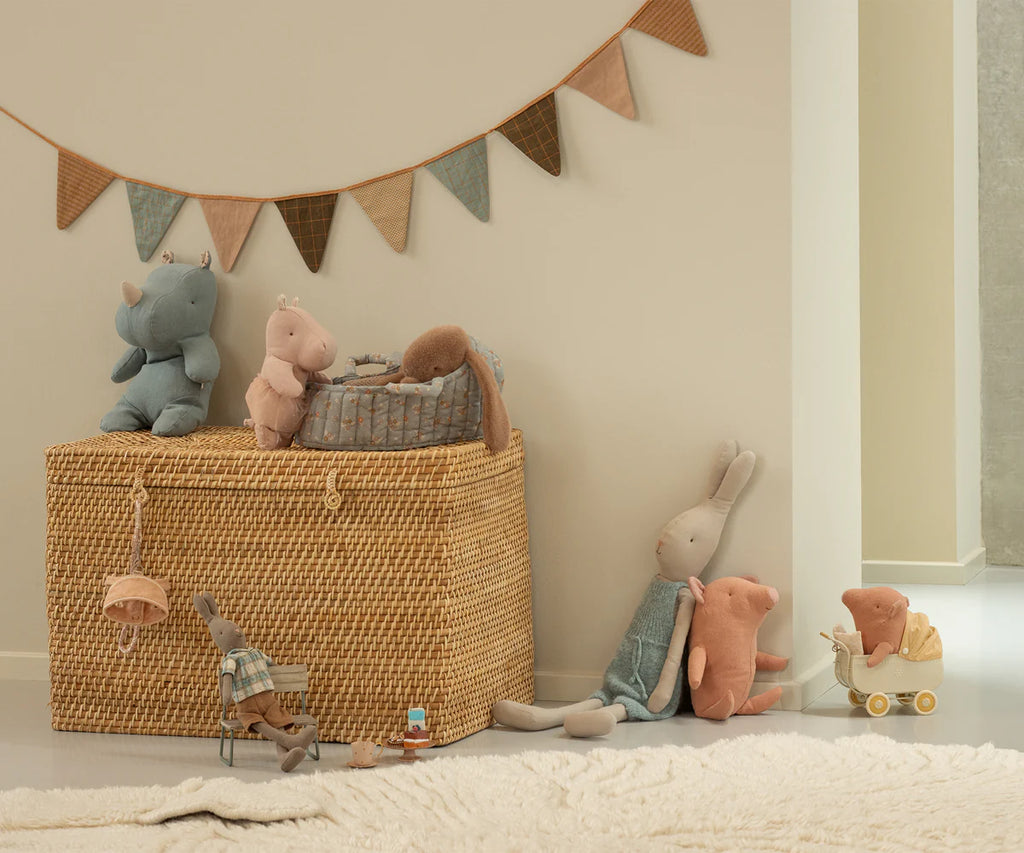 A cozy nursery features the Maileg Small Hippo With Skirt Stuffed Animal - Soft Rose on a woven basket, alongside a stroller toy and soft bunting above a fluffy white rug. The decor is neutral and softly toned.