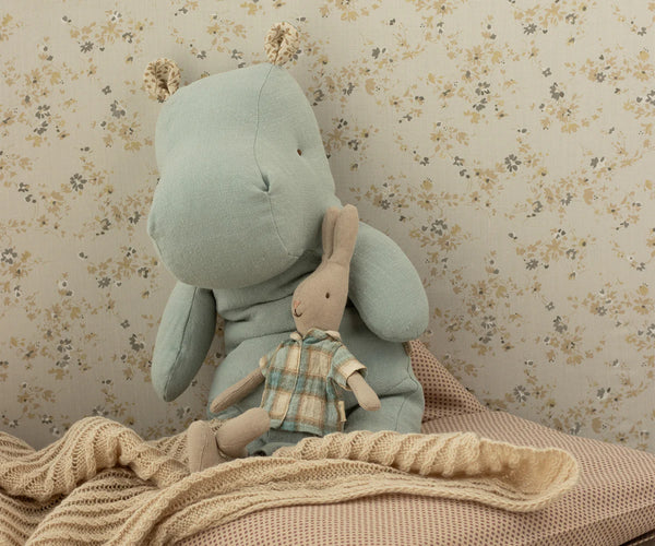 The Maileg Medium Hippo Stuffed Animal in blue sits next to a small rabbit doll in a plaid shirt on a beige knitted blanket against floral wallpaper—adorable safari friends for any cozy space.