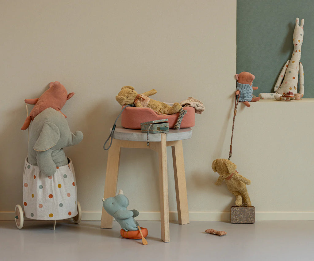 The Maileg Puppy Supply Accessories - Rose features a soft plush dog in a cozy bed atop a stool, with other charming animal toys arranged nearby in a minimal, pastel room for a whimsical, playful display.