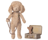 A soft Maileg plush dog with floppy ears stands upright, sporting a collar and leash. Next to it is a small metal suitcase with a Maileg print, containing Puppy Supply Accessories - Rose: a polka-dot bone toy and another cute item.