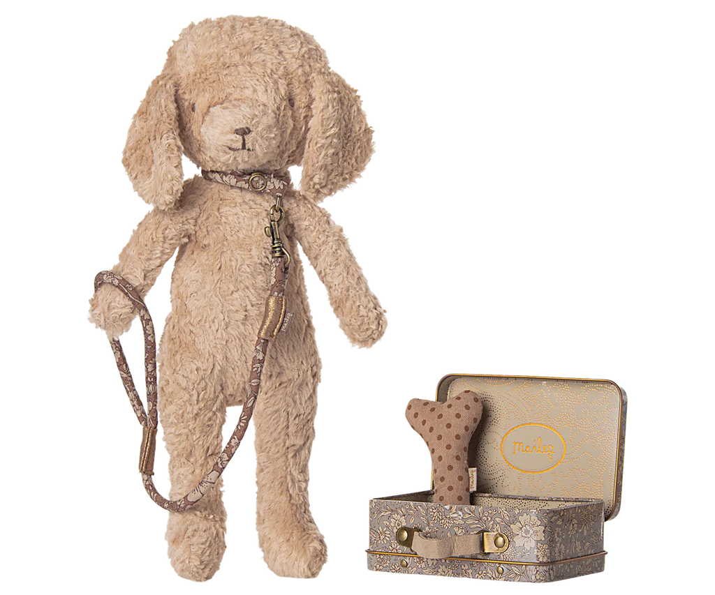 A soft Maileg plush dog with floppy ears stands upright, sporting a collar and leash. Next to it is a small metal suitcase with a Maileg print, containing Puppy Supply Accessories - Rose: a polka-dot bone toy and another cute item.