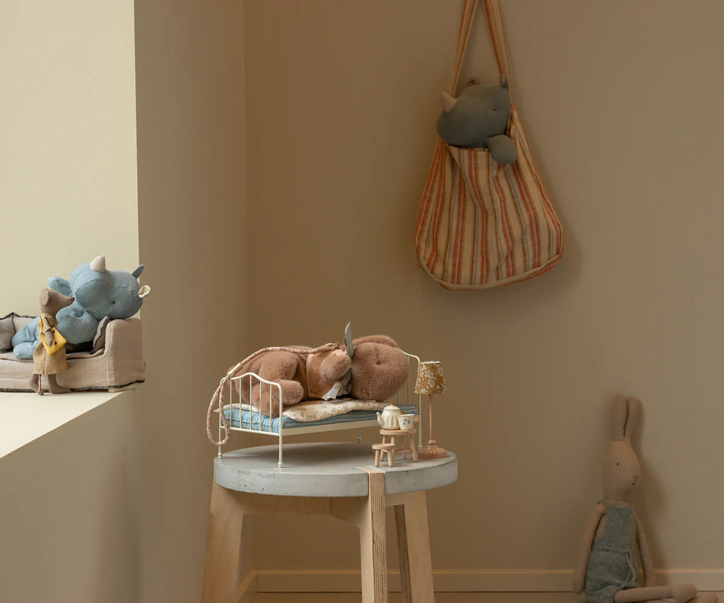 A cozy children's room features stuffed toys: a blue rhino and bunny on a sofa, brown rhino on a miniature bed, blue rhino in a striped bag on the wall, and the Maileg Sleeping Bunny Supply wearing a cute harness sitting against the wall.