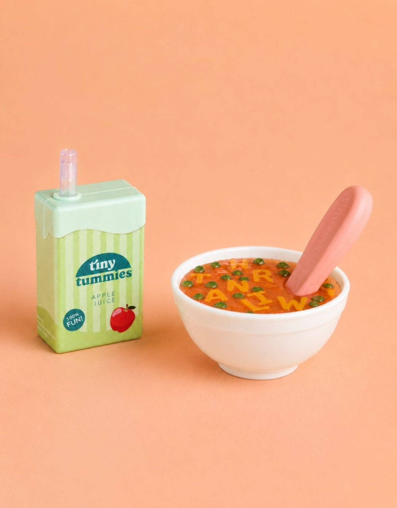 The Tiny Tummies Set - Alphabet Soup Bowl and Juice Box For Dolls features a small juice box and a white bowl of alphabet soup with a pink spoon, all arranged on a peach-colored background, creating the perfect play set for role-playing games or as charming accessories for dolls.