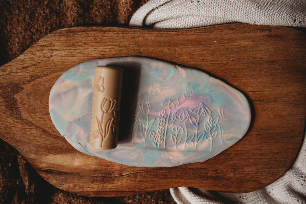 The Flower Roller - Eco Pattern Playdough Roller features elegant floral engravings and rests atop a marbled pastel clay slab imprinted with flower designs, all displayed on a wooden board with a textured cloth nearby—perfect for sensory play and inspiring creative adventures with eco-friendly playdough tools.