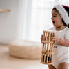 A toddler wearing a white onesie and a Santa hat is smiling while holding the Wooden Rain Maker, a beautifully crafted toy made from non-toxic materials. The background is softly lit with natural light, revealing a blurred wicker chair in a neutral-colored room.