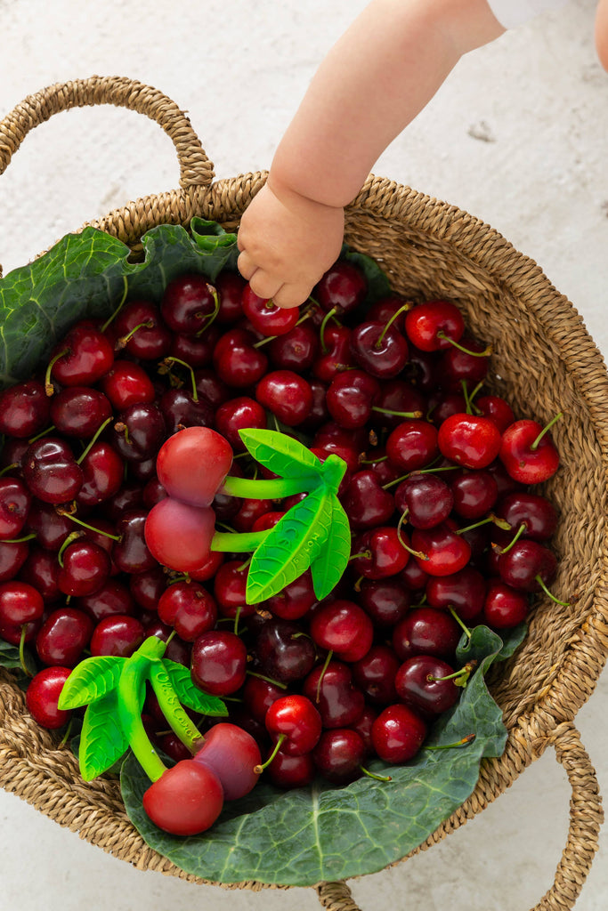 A child’s hand reaches into a woven basket of ripe cherries—some are actually Mery the Cherry Teether baby toys, made from natural rubber with green plastic leaves—resting on green leaves against a light background.