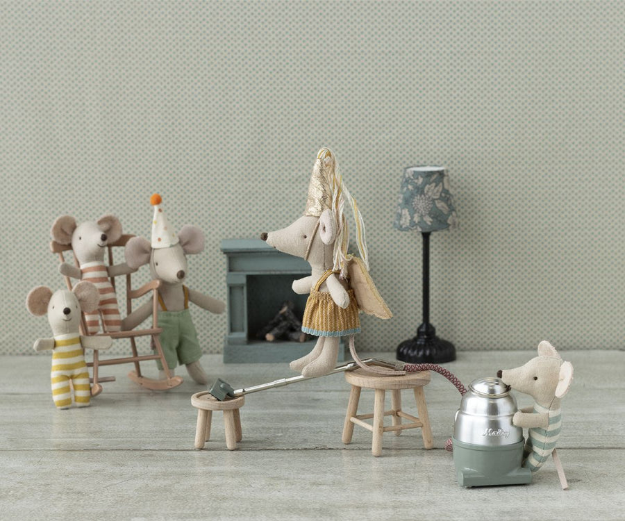 A group of toy mice are having a whimsical tea party. One little brother mouse vacuums while another sits on a stool in Maileg Clown Clothes In Suitcase, Little Brother Mouse. A third mouse stands on stilts wearing a cone hat, and two mice on rocking chairs look on. Nearby, a small metal suitcase sits beside them against the muted, patterned wallpaper.