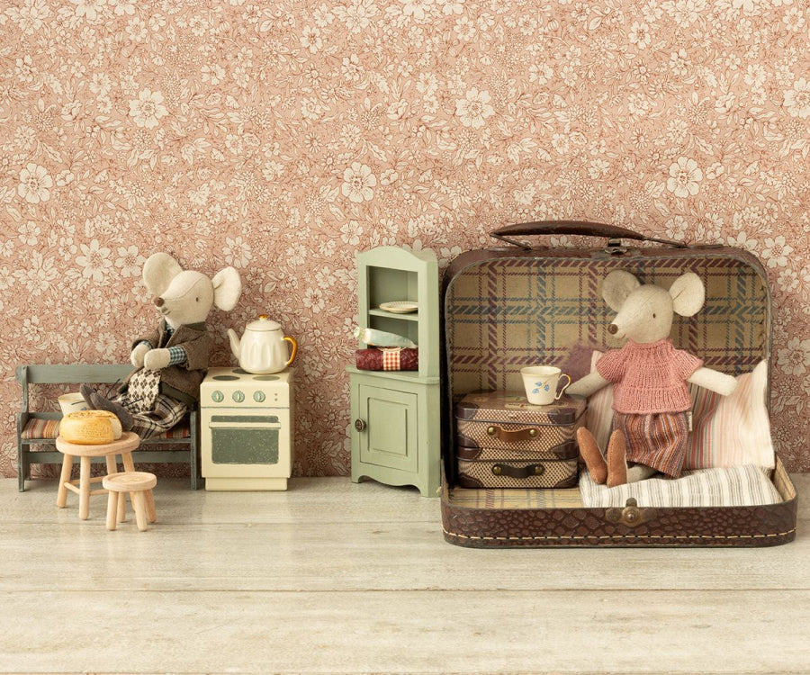 A whimsical scene with toy mice in a vintage-style setup. One mouse, dressed in the Maileg Knitted Blouse And Skirt In Suitcase, Grandma Mouse clothes, sits in an open metal suitcase. Another, in a dress, stands near a small stove. Accessories include a tiny teapot, cup, and a small wooden table with stools, all against a floral backdrop.