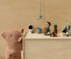 A soft brown teddy bear sits by a wooden box topped with miniatures: a toilet, watering can, blue bucket, Maileg Cowboy Mouse On Rocking Horse (Little brother), tiny cactus, and tent. The background is plain light wood. Ships in one week.