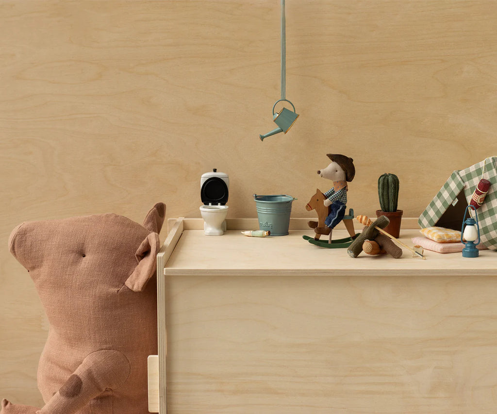 A soft brown teddy bear sits by a wooden box topped with miniatures: a toilet, watering can, blue bucket, Maileg Cowboy Mouse On Rocking Horse (Little brother), tiny cactus, and tent. The background is plain light wood. Ships in one week.