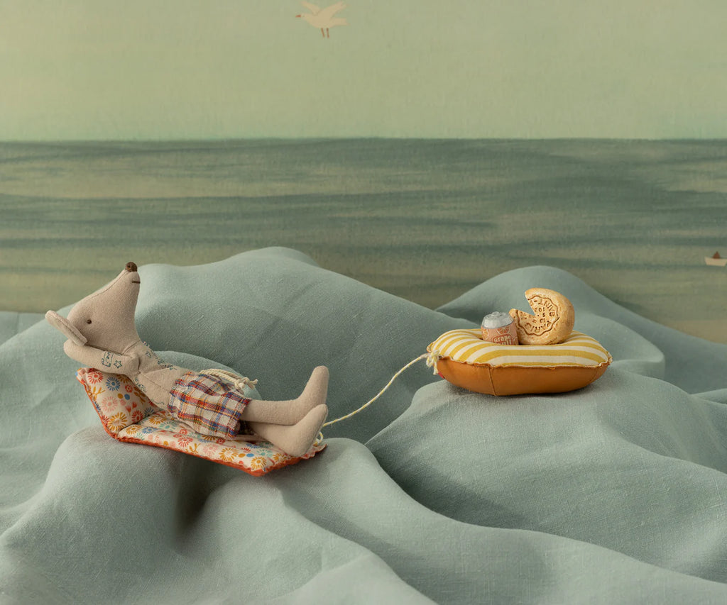 The Maileg Beach Mouse with Air Mattress - Dad lounges in plaid shorts on a blue air mattress, relaxing by an illustrated summer sea scene. Nearby, a yellow raft with a toy and cookie floats, tethered by string.