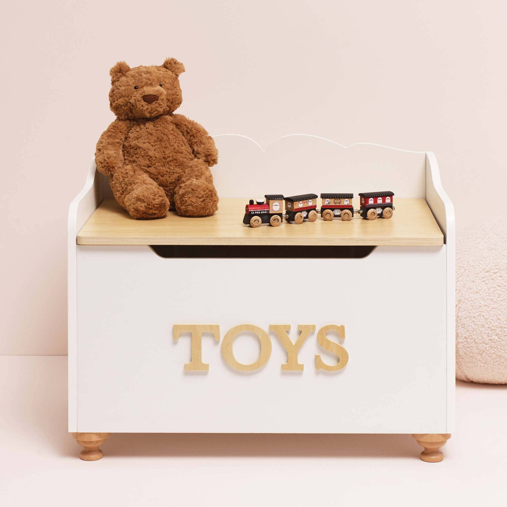 The Toy Storage Box, labeled "TOYS," is white and displayed with a brown teddy bear and a wooden toy train on top, all set against a soft pink background.