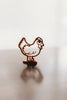 A brown chicken-shaped cutter from the Farm Cutter Set - Eco Playdough Tool collection stands upright on a reflective surface against a blurred white background.