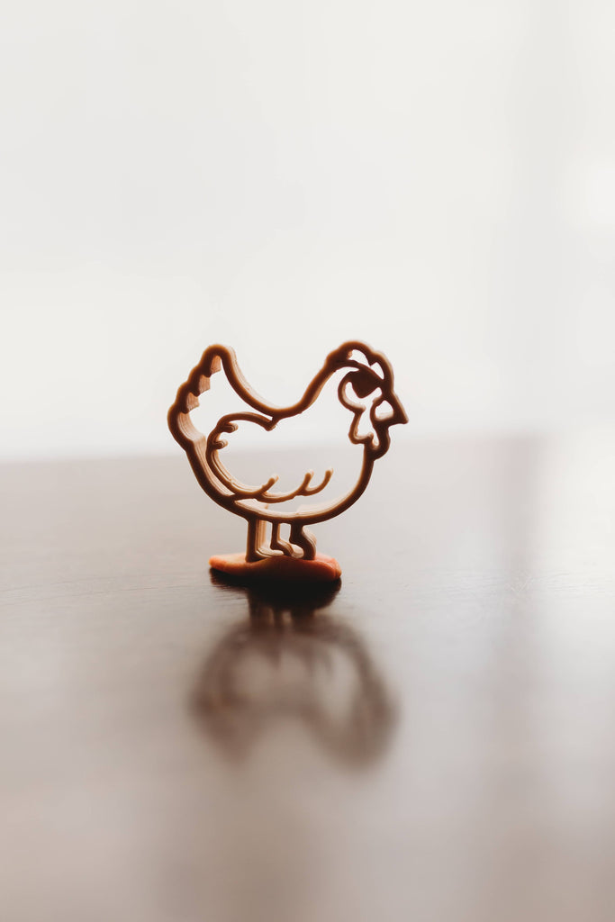 A brown chicken-shaped cutter from the Farm Cutter Set - Eco Playdough Tool collection stands upright on a reflective surface against a blurred white background.
