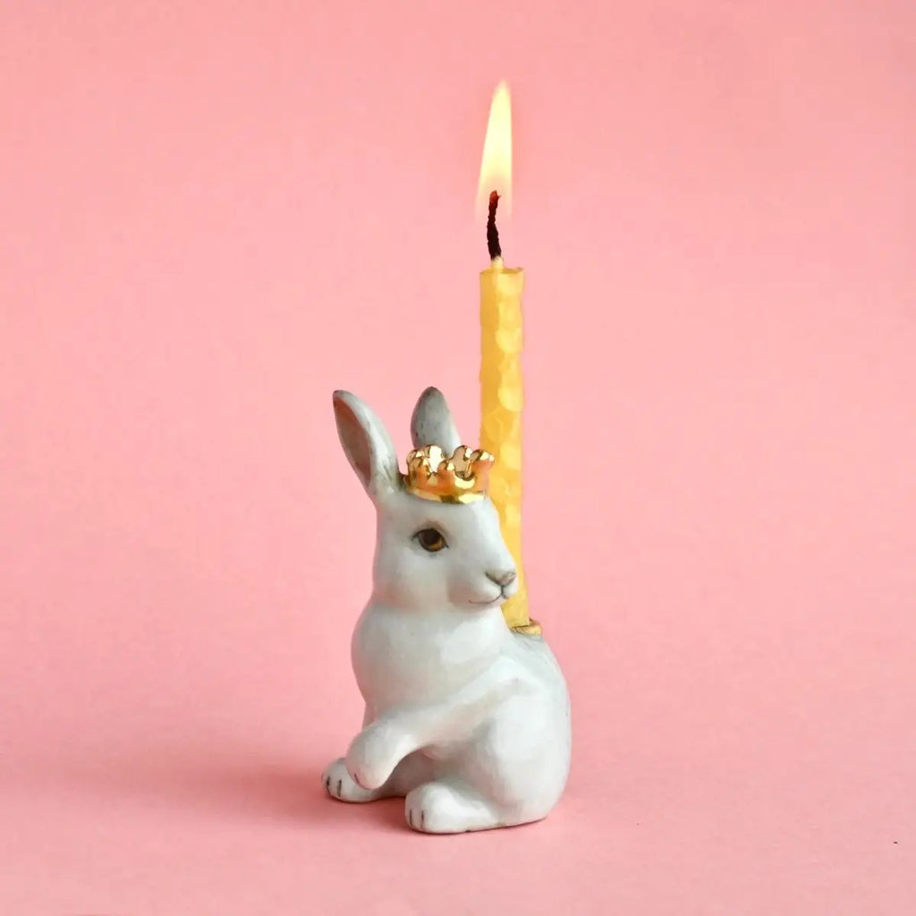 The Royal White Rabbit Cake Topper, a ceramic figurine with a golden crown, holding a lit yellow candle on its head, set against a soft pink background. This hand-painted porcelain piece is an heirloom quality.