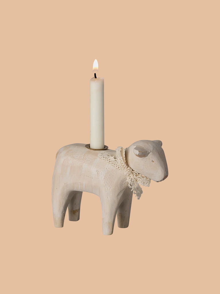 An Easter table is graced by the Maileg Wooden Candle holder, shaped like an off-white lamb, and adorned with a lace ribbon around its neck. A white candle flickers gently on its back, set against a plain beige background, adding warmth and charm to the scene.