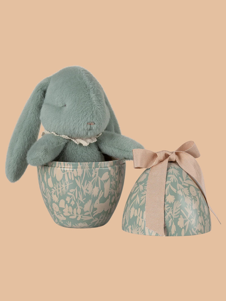 The PRE-ORDER: Maileg Easter Egg With Bunny - Mint features a soft green plush rabbit inside a floral-pattern half-egg container, with the other half, decorated with a beige ribbon, resting beside it against a soft peach background. Ships in February.