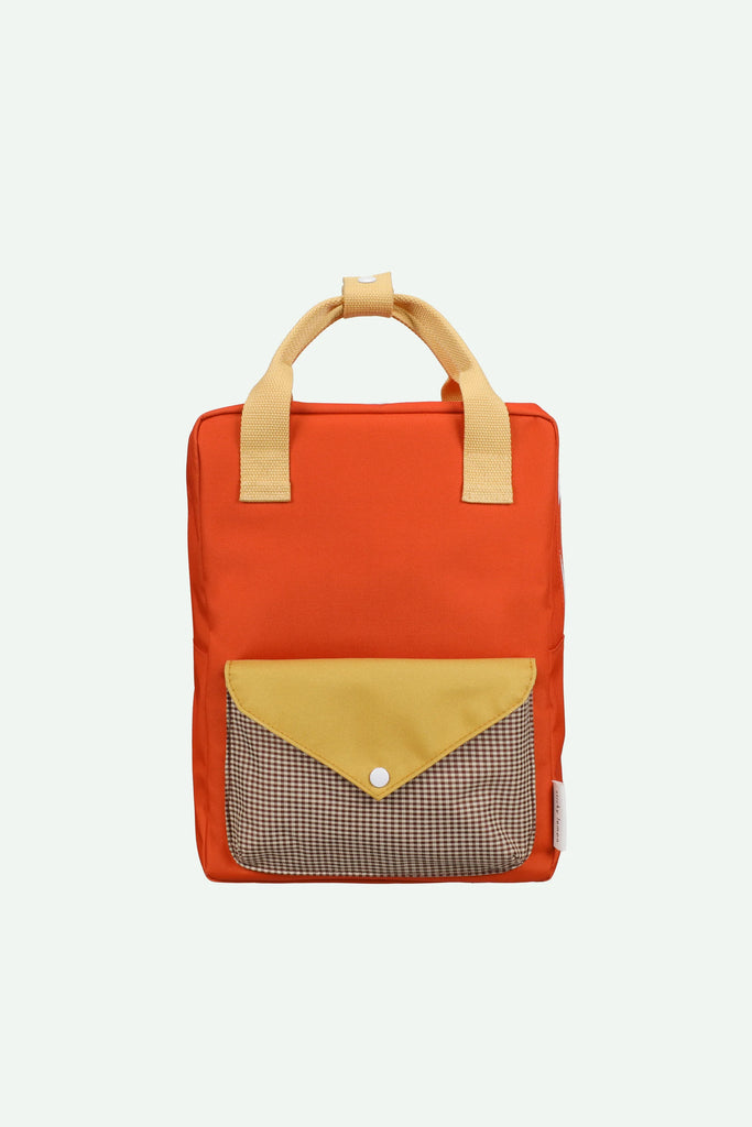 A Sticky Lemon Backpack Medium | Envelope | Campfire Orange is shown: a medium-sized, campfire orange backpack with beige handles and a front pocket. The front pocket has a yellow flap with a button and a brown checkered section underneath. Made from recycled PET material with a waterproof lining, this backpack is both stylish and eco-friendly. The background is white.