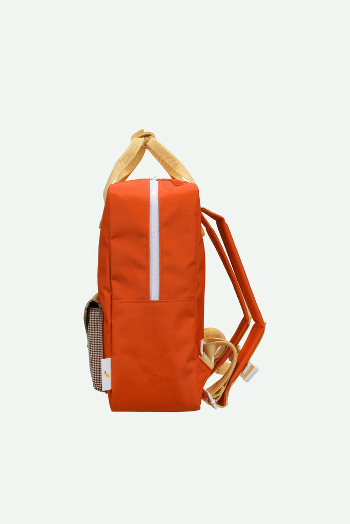 The Sticky Lemon Backpack Medium | Envelope | Campfire Orange is shown from the side, featuring a campfire orange color with beige straps and a white zipper running vertically on the front. Crafted from recycled PET bottles and finished with a waterproof lining, this backpack offers both sustainability and style. The background is plain and light-colored.