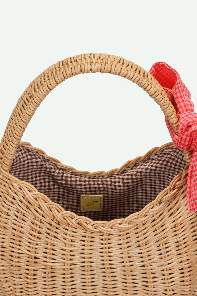 The Sticky Lemon Wicker Basket | Lemon Tree is a delightful kids accessory crafted from woven straw, featuring a rounded handle, brown checkered fabric lining, and a playful red gingham bow tied to one side—evoking the charm of a classic wicker basket and adding a whimsical touch to any outing.