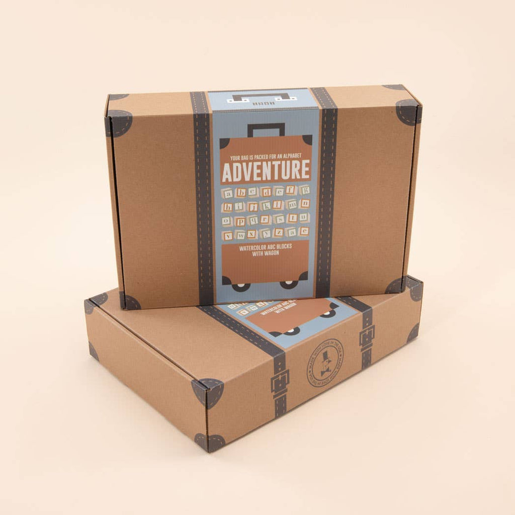 Two brown cardboard boxes styled as vintage suitcases with "ADVENTURE" graphics are stacked beside the Uncle Goose Watercolor ABC Blocks with Wagon, all set against a neutral background.