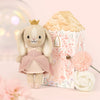 Cuddle + Kind Tiny Rose the Princess, a fair trade hand-knit doll in a pink dress and gold crown, stands beside a pastel house-shaped gift box with pink and white flowers on a soft pink background.