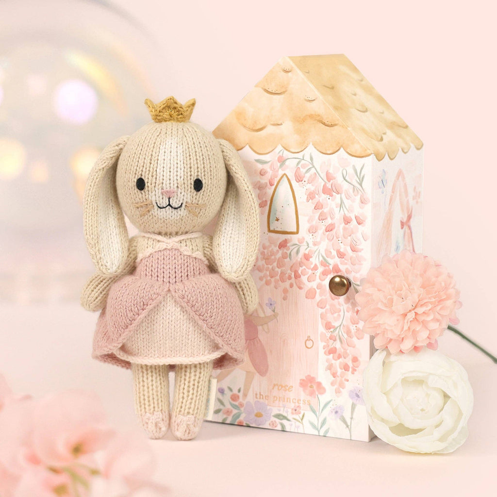 Cuddle + Kind Tiny Rose the Princess, a fair trade hand-knit doll in a pink dress and gold crown, stands beside a pastel house-shaped gift box with pink and white flowers on a soft pink background.