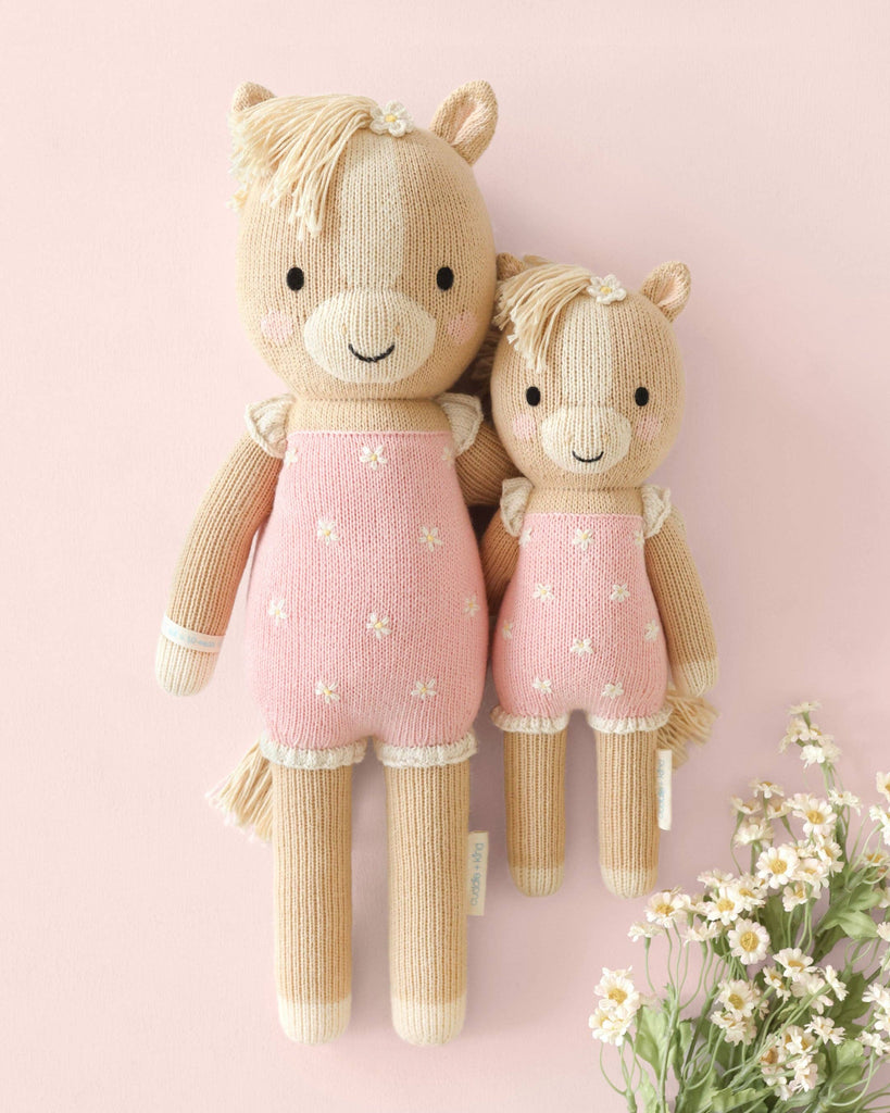 Two Cuddle + Kind Sophia the Pony dolls, with beige bodies and manes, wear matching pink floral dresses on a pastel pink background beside white daisies. These fair trade treasures are handmade with love in Peru.