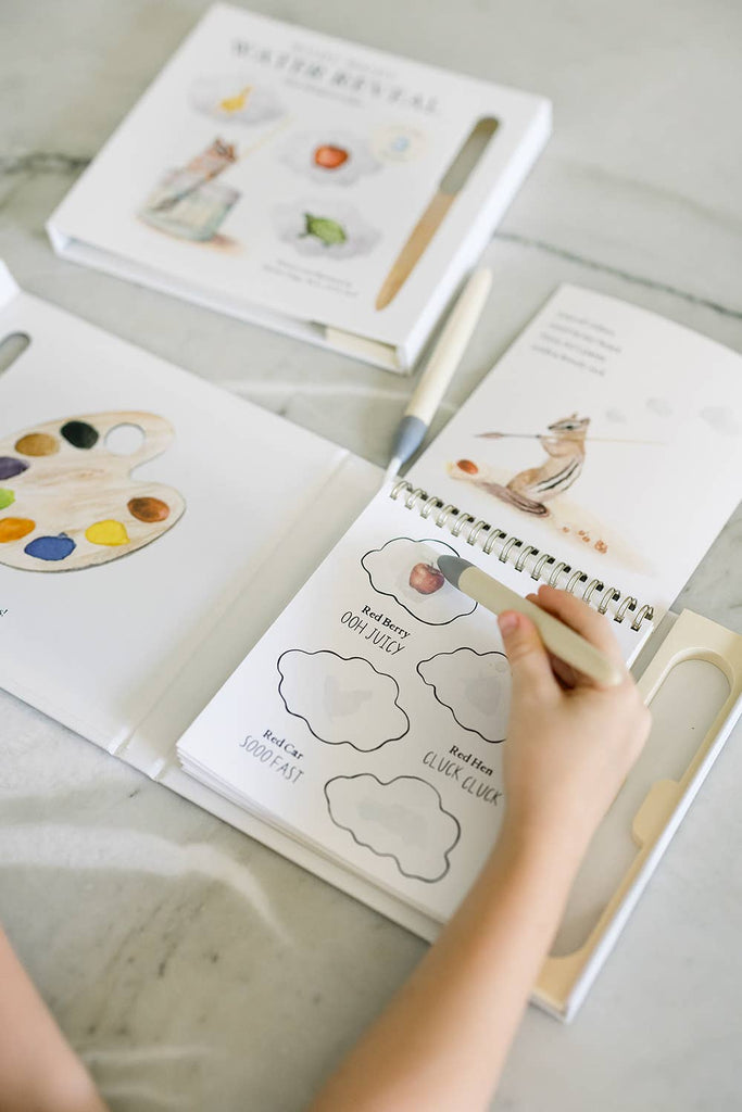 A child holds a pencil over an open Wishy Washy (Water Reveal Edition) activity book with cloud-shaped writing spaces—perfect as an early learning gift. Nearby are illustrated books and a paint set on a light-colored table.