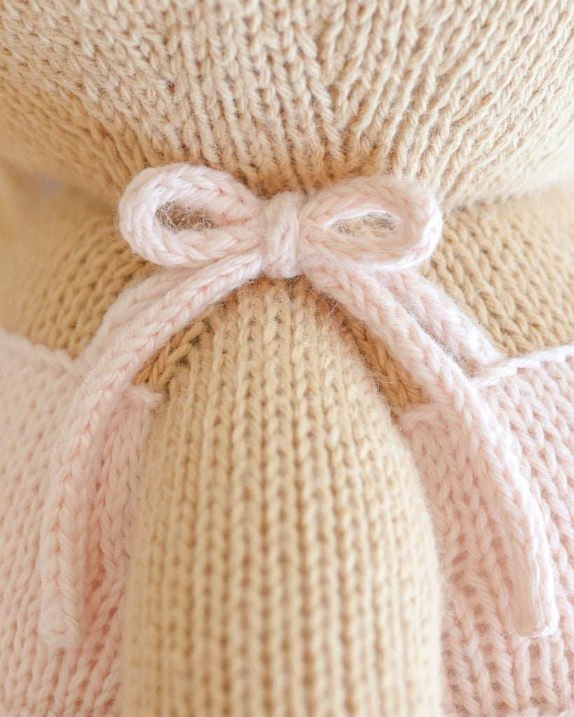 Close-up of a light pink knitted bow tied around the back of Cuddle + Kind Kelsie the Pony, a beige, hand-knit doll. Detailed yarn textures highlight this charming creation, handmade in Peru.