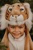 A young child wearing the Costume - Tiger, a plush faux fur tiger hat and white sweater, smiles at the camera amid green foliage.