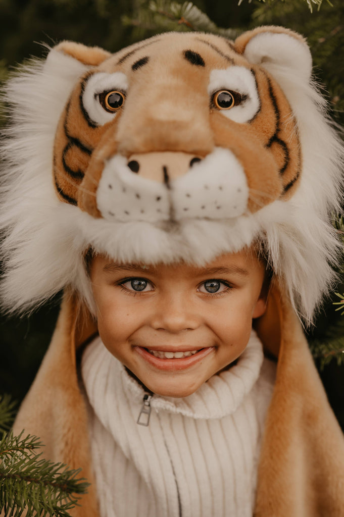 A young child wearing the Costume - Tiger, a plush faux fur tiger hat and white sweater, smiles at the camera amid green foliage.
