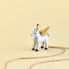 A small, white porcelain figurine of a winged unicorn with golden wings, standing next to a delicate Gold Pegasus Necklace on a plain beige background.