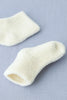 A pair of small, white, fluffy Baby Pile Knit Socks - Natural are placed on a light blue fabric surface. One sock is positioned flat, revealing its opening, while the other is slightly rolled, emphasizing its soft and textured material. These adorable Lithuania baby accessories add a touch of cozy charm to any outfit.