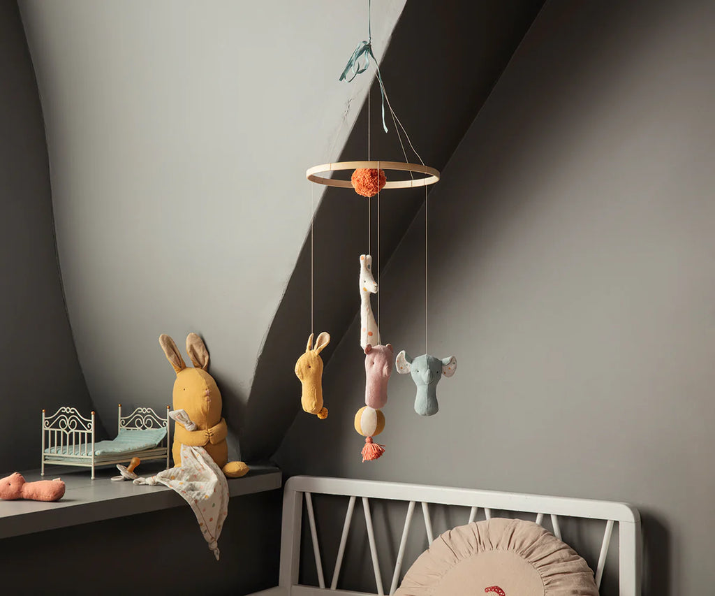 A cozy nursery corner with a soft toy rabbit on a white bench, a small toy bed, and the Maileg Lullaby Friends Mobile featuring pastel animal plushies, set against gray walls with gentle lighting.