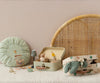 A soft, cozy scene with small suitcases containing plush mice, a Maileg Small Round Cushion in mint, a miniature rocking chair, tiny lamp, mugs, and a wicker headboard against a beige wall.