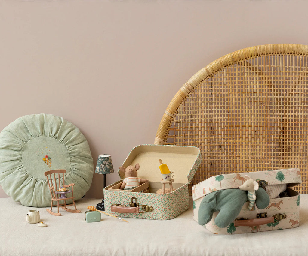 A soft, cozy scene with small suitcases containing plush mice, a Maileg Small Round Cushion in mint, a miniature rocking chair, tiny lamp, mugs, and a wicker headboard against a beige wall.