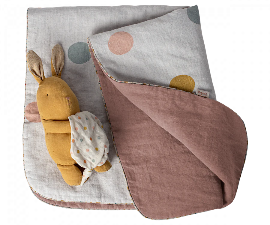 A mustard-yellow plush rabbit toy with long ears and a small cloth backpack rests on the Maileg Small Blanket - Rose. This cozy nursery essential features pastel-colored polka dots on one side and plain mauve, crafted from soft linen fabric, on the other.