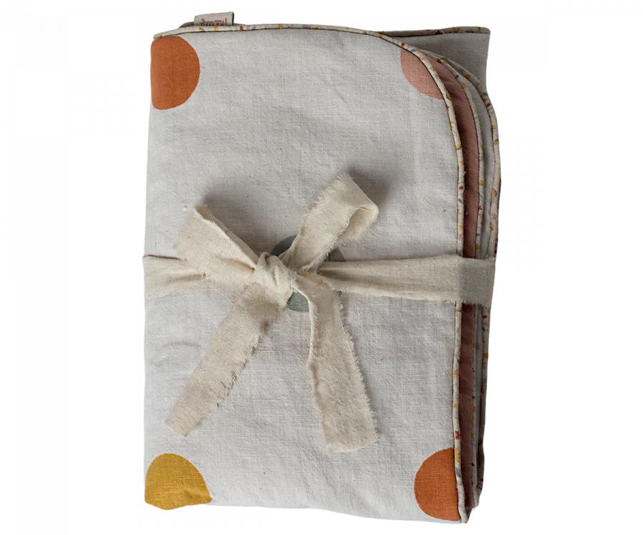 A Maileg Small Blanket - Rose, folded and bound with a natural-colored ribbon, features white linen fabric adorned with orange, yellow, and brown circular patterns, making it perfect for a nursery.