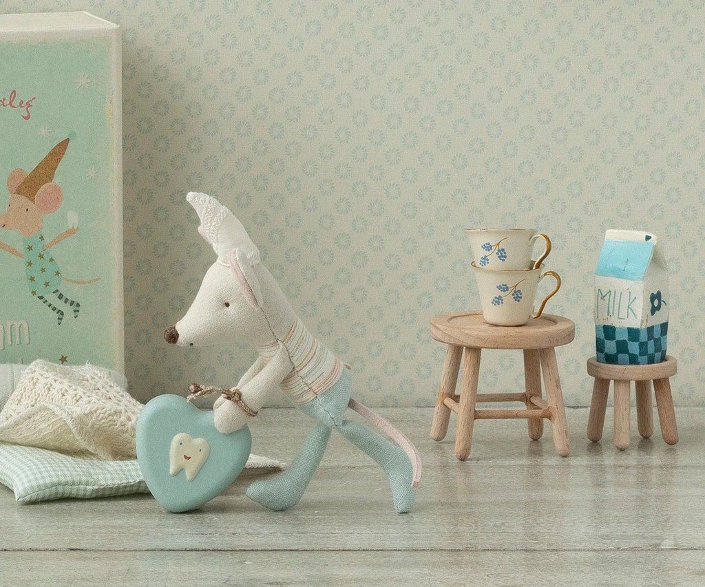 A plush toy mouse, dressed in light blue overalls, carries a blue heart-shaped box with a tooth illustration. Behind, the Maileg Table & Stool Set, Mouse features stacked teacups and a milk carton, all set against pastel-patterned wallpaper that complements the cozy charm of the Mouse family furniture.