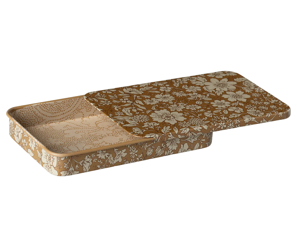 The Maileg Metal Box with Sliding Lid is a rectangular decorative tray featuring a detachable lid adorned with intricate white floral patterns on a brown background. The lid is partially slid open to the side, revealing the matching interior design inside. This storage box exudes a vintage and elegant appeal.