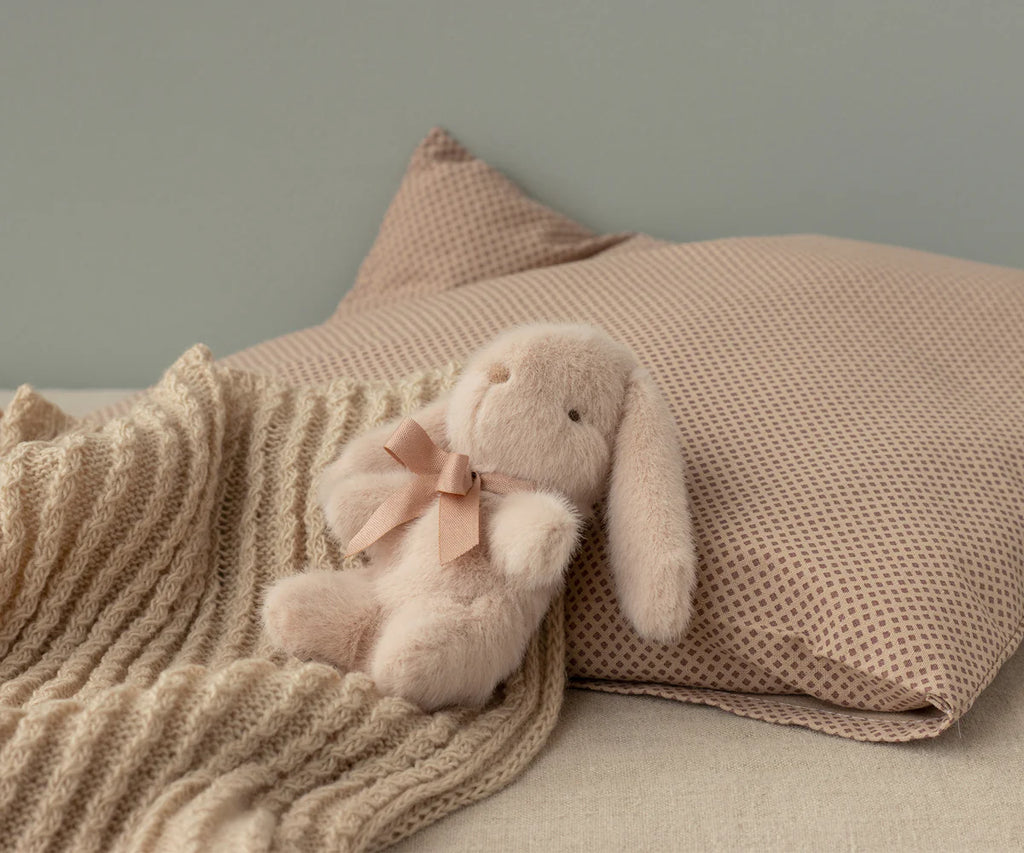 A soft, beige stuffed bunny with a pink bow rests beside the Maileg Baby Blanket and a textured beige pillow on a neutral-colored bed.