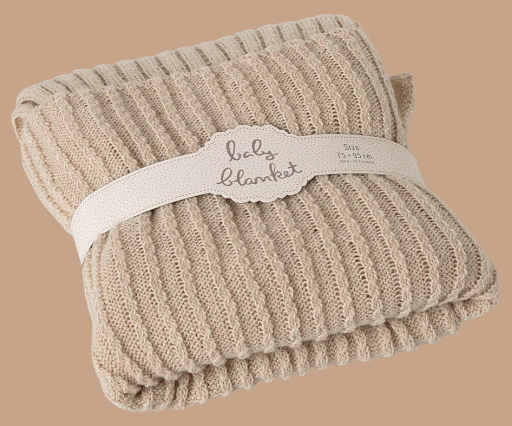 The Newborn Gift Set - Natural includes a folded beige ribbed knit baby blanket crafted from natural Peruvian cotton yarn, wrapped with a white "baby blanket" band, and shown against a light brown background.