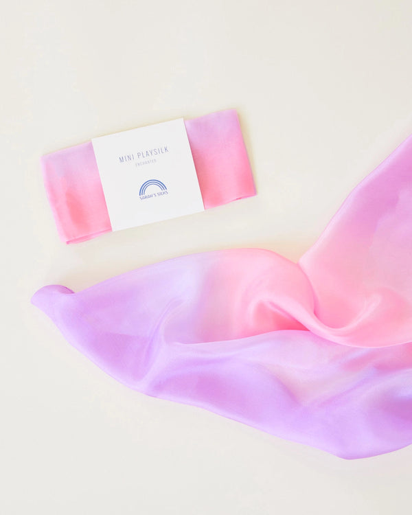 A pastel pink and purple silk scarf gracefully unfolding near a minimalist card labeled "Sarah's Silk Enchanted Playsilk - Blossom" in a light, soft cream background, ideal for imaginative play.