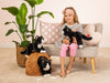 A young girl wearing a striped dress and pink leggings is sitting on a beige sofa, holding a Teddy Hermann Black Cat Stuffed Animal. Two other similar plush cats are placed in a wicker basket and inside a small tunnel on the floor. A plant and decorative pillows are beside her.