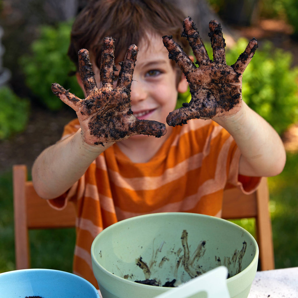 A young child in an orange striped shirt smiles, holding up soil-covered hands while sitting outdoors at a table with bowls of dirt—enjoying the DIY Seed Ball Kit - Rainbow Garden STEM activity with non-GMO seeds.