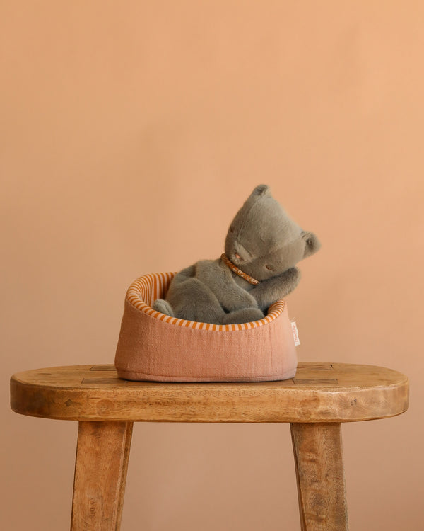 The Maileg Small Sleeping Kitten in Basket, featuring a gray plush kitten with a beaded collar, rests in a small peach basket atop a wooden stool against a plain peach background.