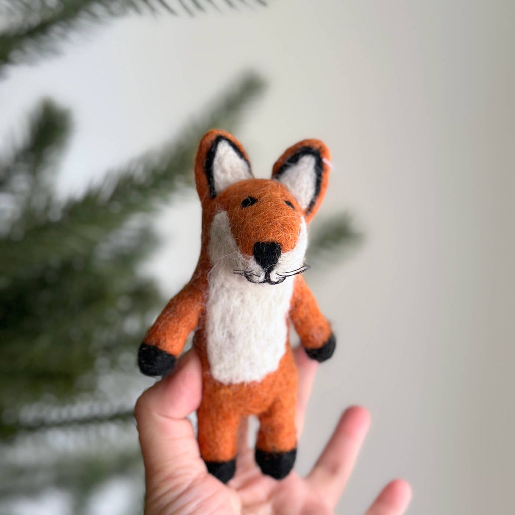 A hand holds the Felt Finger Puppet: Fox, a small handmade wool puppet with white, orange, and black details—ideal for storytelling. A blurred green pine tree appears in the background.