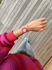 A person in a pink and red sweater, light blue jeans, and blue socks stands on wooden flooring, showing their left wrist wearing the Mini Kyomo Watch - Disco. The photo is taken from above.